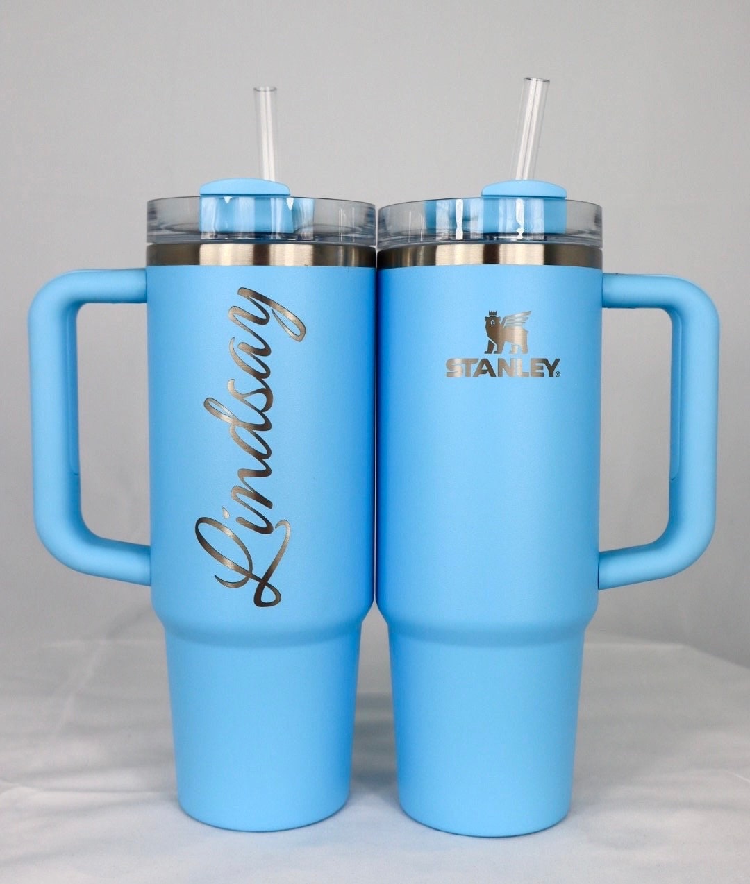 Two pale blue Stanley tumblers, one engraved with a name running vertically up the back in a script font