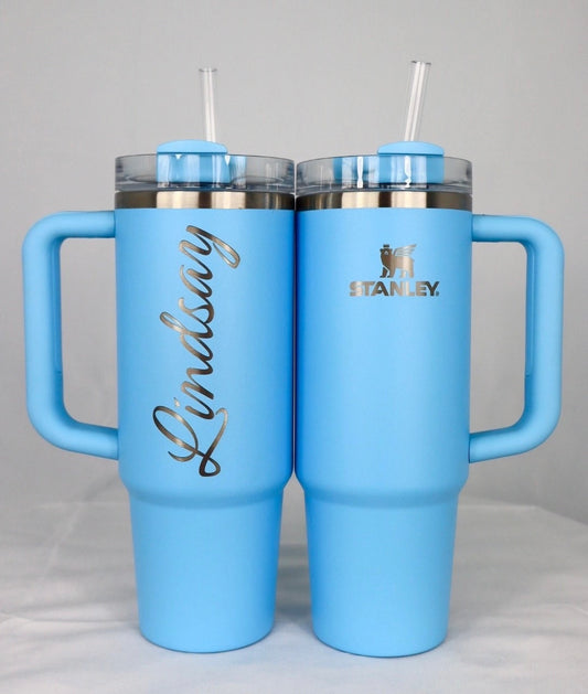 Two pale blue Stanley tumblers, one engraved with a name running vertically up the back in a script font