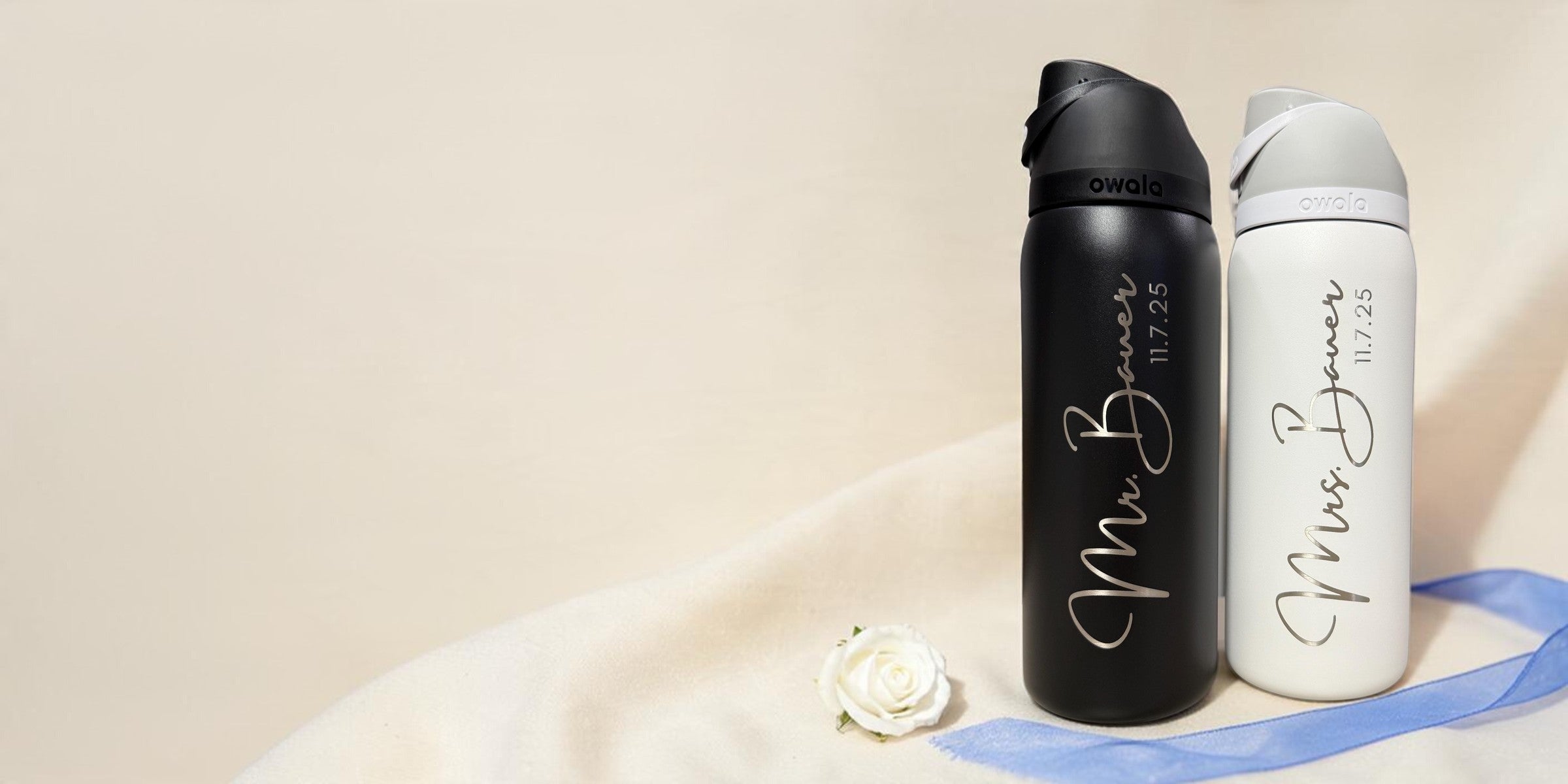 A black and a white Owala FreeSip engraved Mr. Bauer and Mrs. Bauer, 11.7.25, with a pale blue wedding ribbon